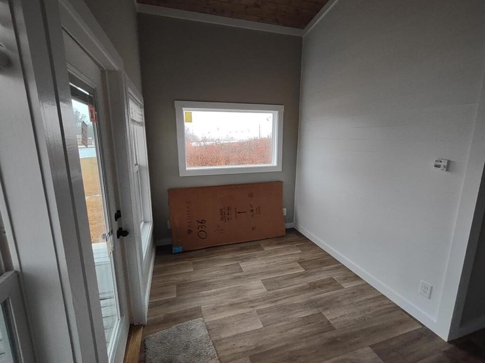 Small room with light gray walls, wooden floor, and a large window showing an outdoor view. A rectangular cardboard piece leans against the wall, conveying an unfinished or transitional space.