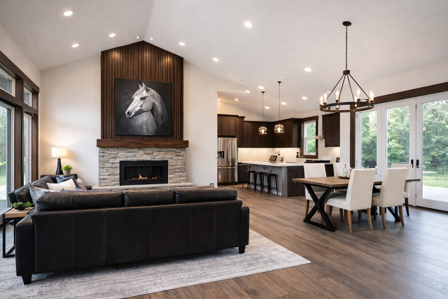 Spacious living area with a dark leather sofa facing a stone fireplace, above which is a horse portrait. Modern kitchen and dining area with large windows and wooded view.