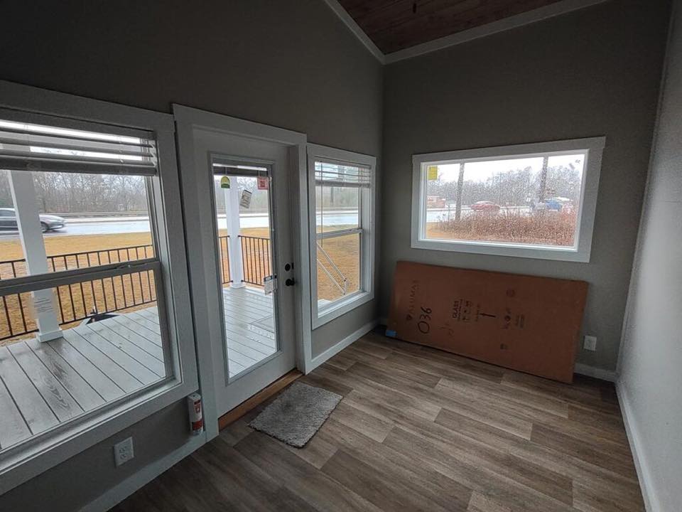 Small room with light wood flooring, large windows, and a glass door leading to a porch. An overcast day is visible outside, conveying a serene mood.