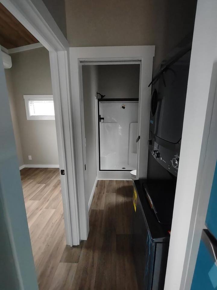 Narrow hallway in a modern home with wood floors, a white shower stall at the end, and stacked washer and dryer on the right, creating a functional space.