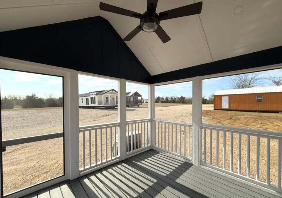 View from a screened porch with white railing and a ceiling fan. Outside, there are several small buildings and a gravel lot under a clear sky. Calm atmosphere.
