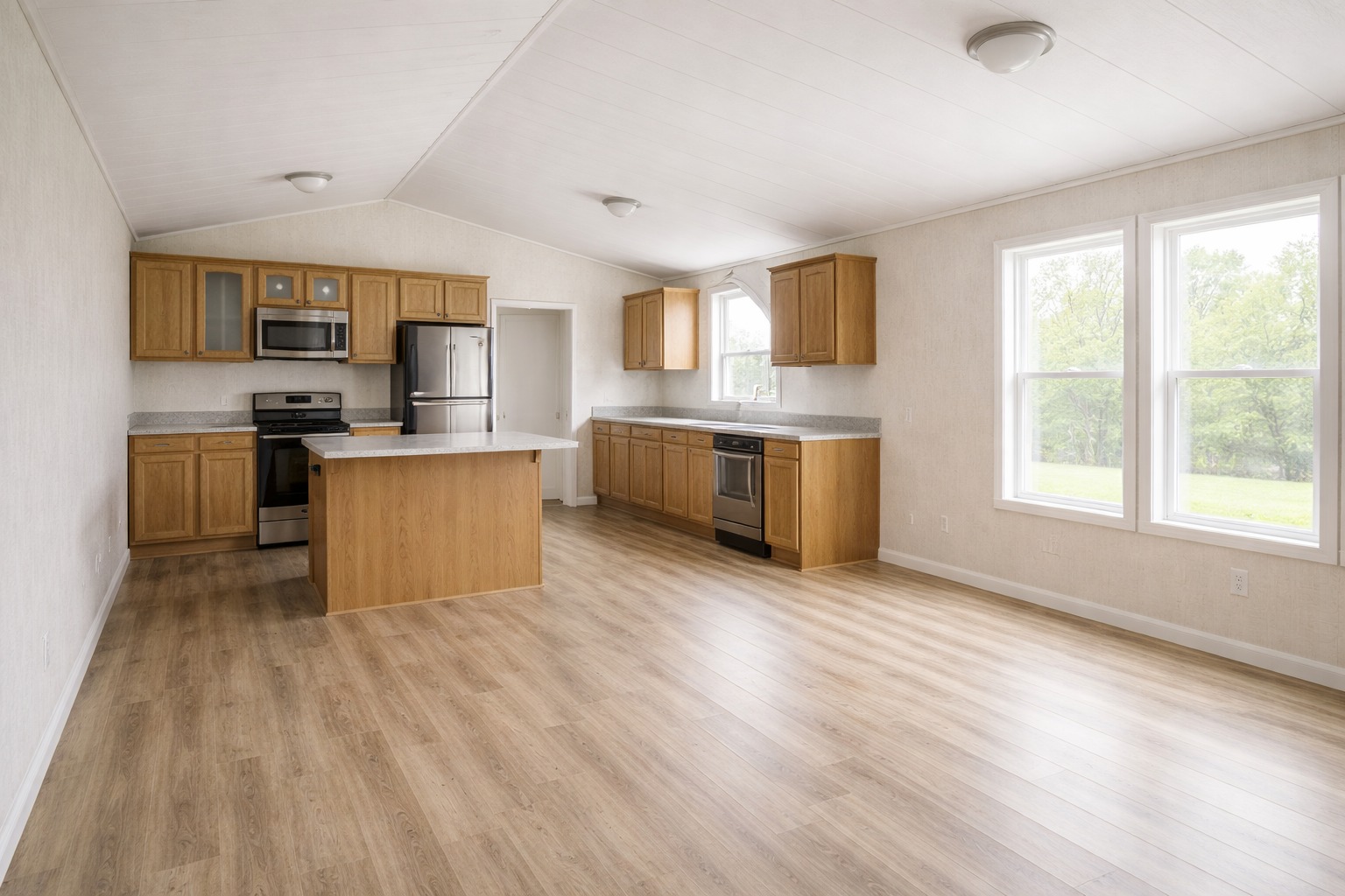 Spacious kitchen with light wood flooring, wooden cabinets, and stainless steel appliances. Large windows to the right offer a serene view of greenery.