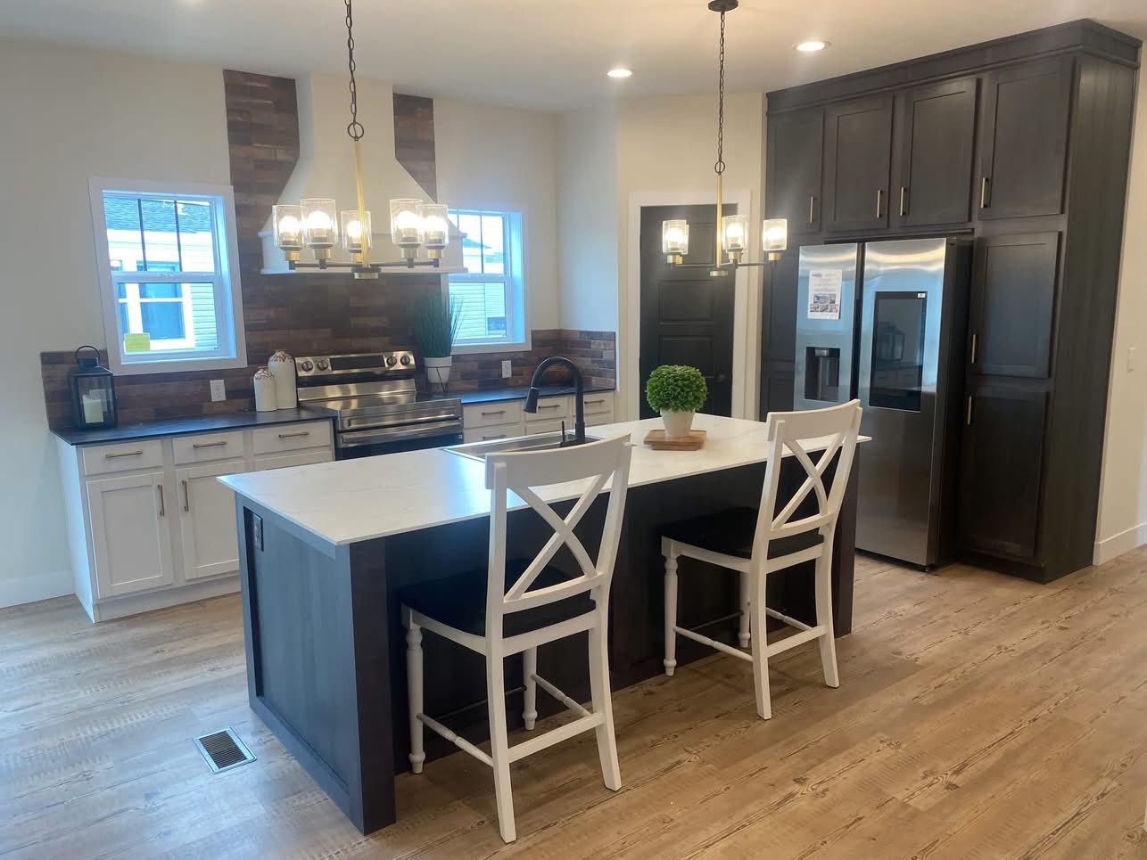 Modern kitchen with a dark island and white countertop, two white chairs, stainless steel appliances, dark cabinets, wood flooring, and bright lighting.