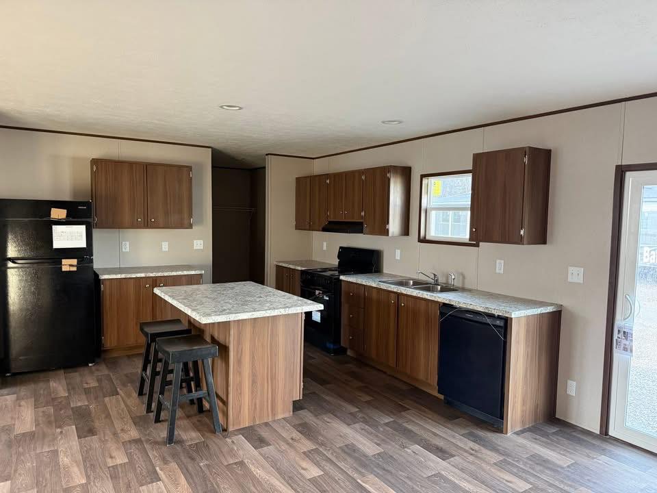 Modern kitchen with wood cabinets, a central island with stools, and dark appliances. The space feels spacious and clean, with a warm, inviting tone.