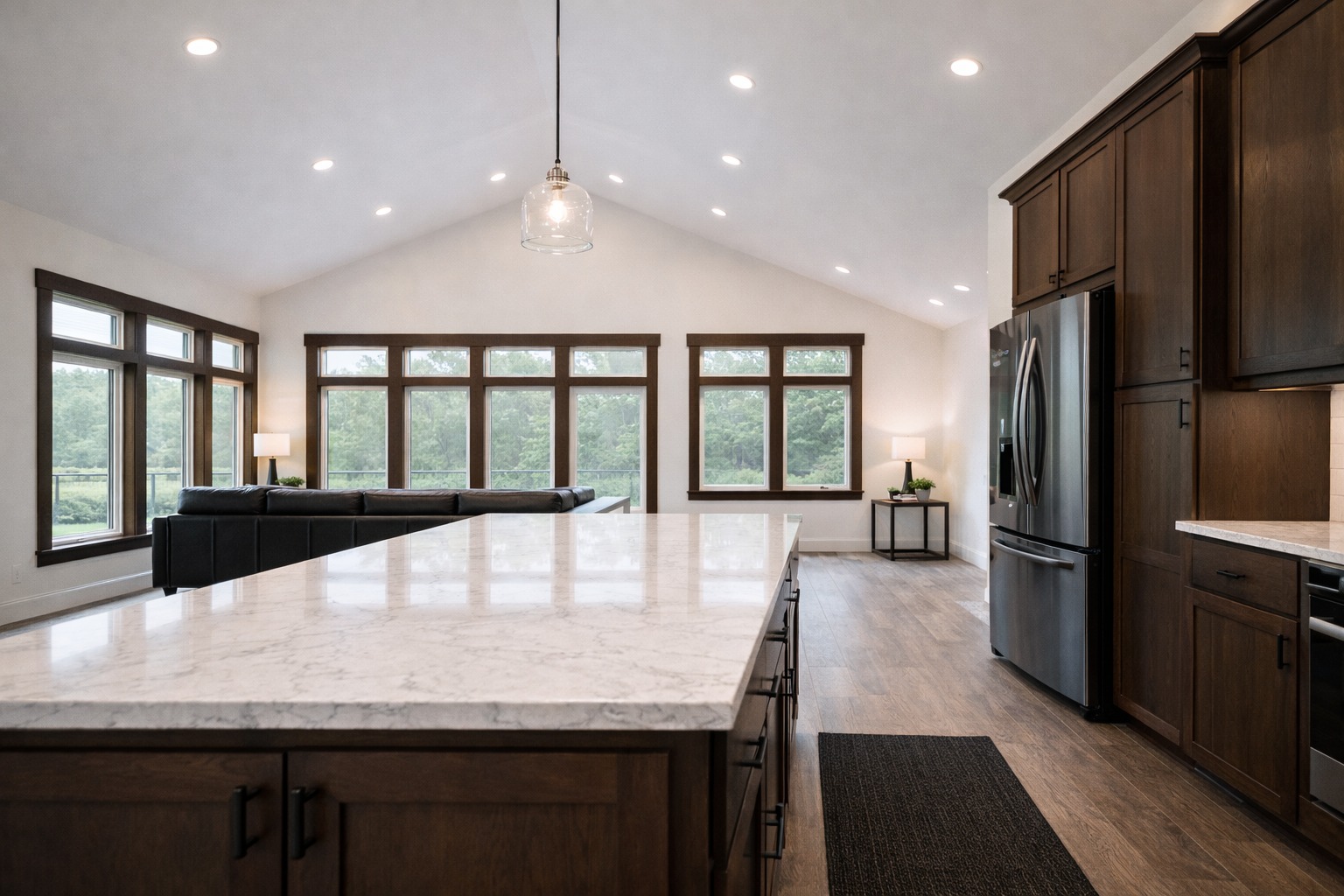 Spacious kitchen with a marble island, dark wood cabinets, and stainless steel appliances. Large windows offer a serene outdoor view, creating a bright and airy ambiance.