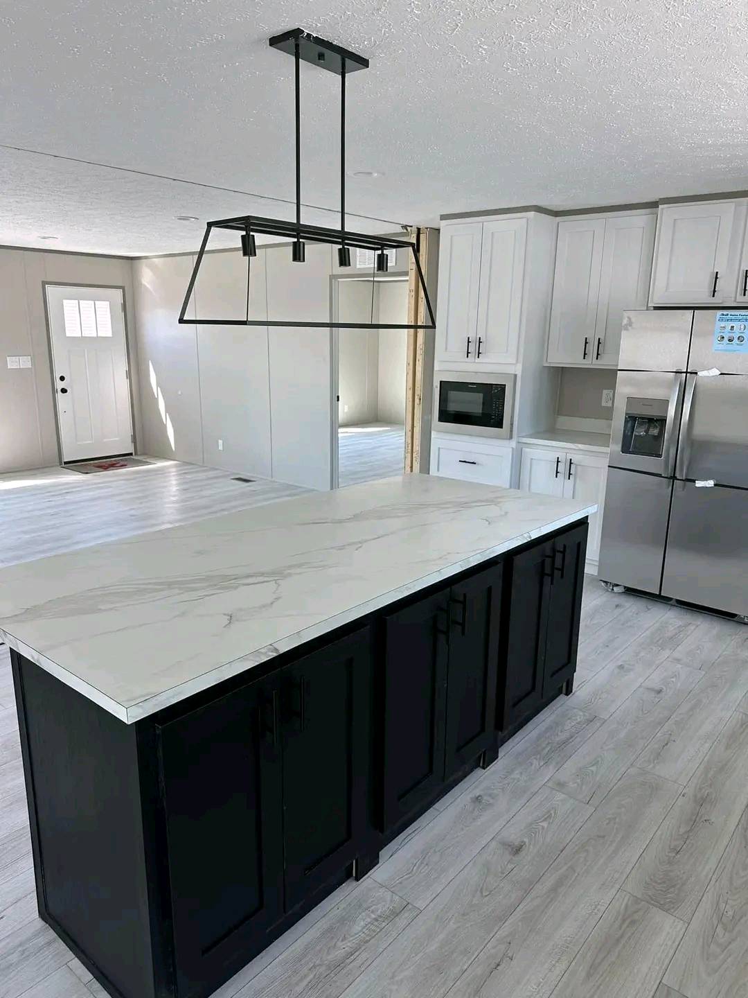 Modern kitchen with a marble countertop island, black cabinets, and light wood flooring. White cabinets, a stainless steel fridge, and a sleek pendant light hang above. Airy and minimalist.