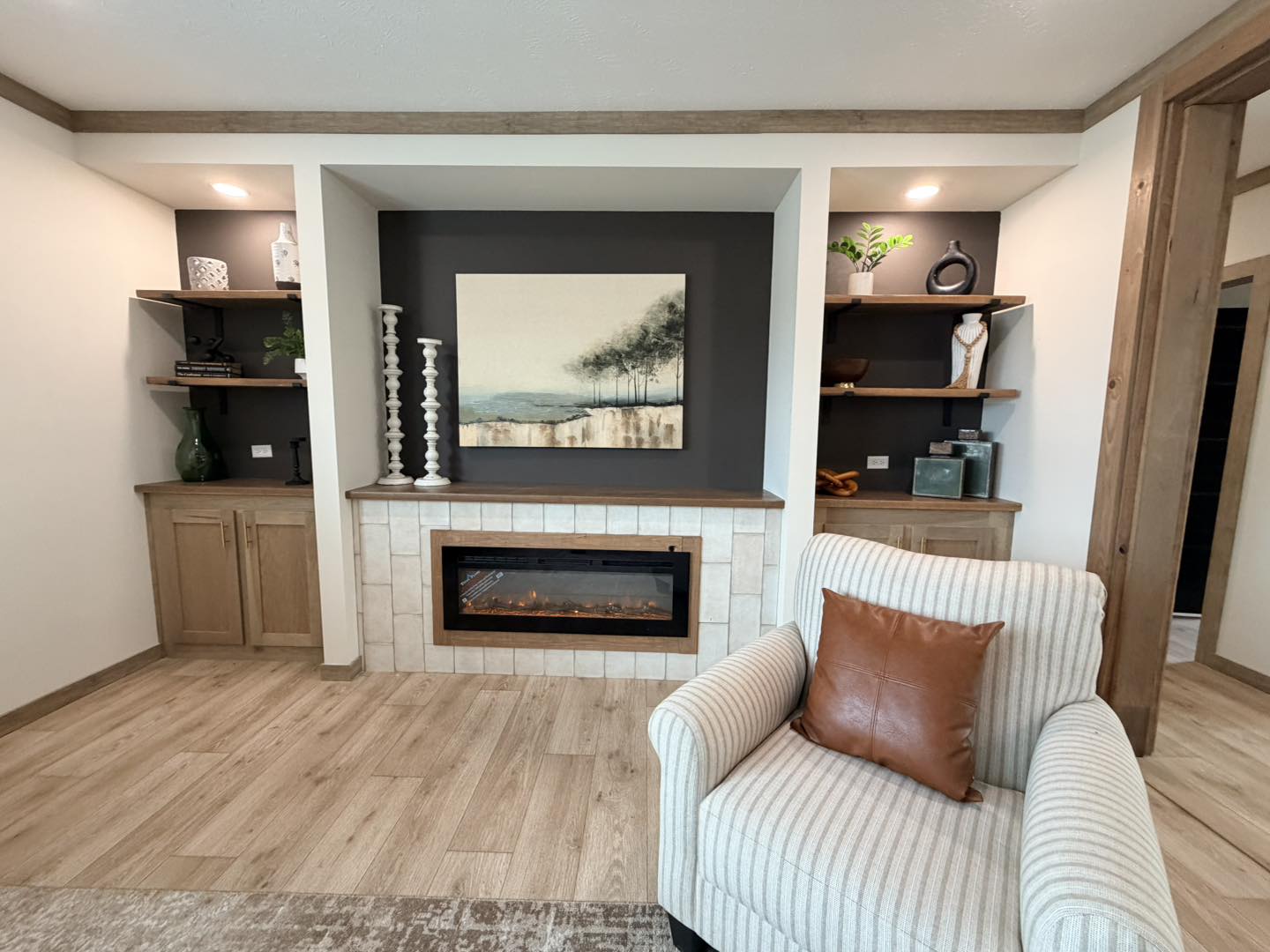 A cozy living room features a modern fireplace, abstract tree artwork, and wood cabinetry. Shelves display decorative items. Warm, inviting ambiance.