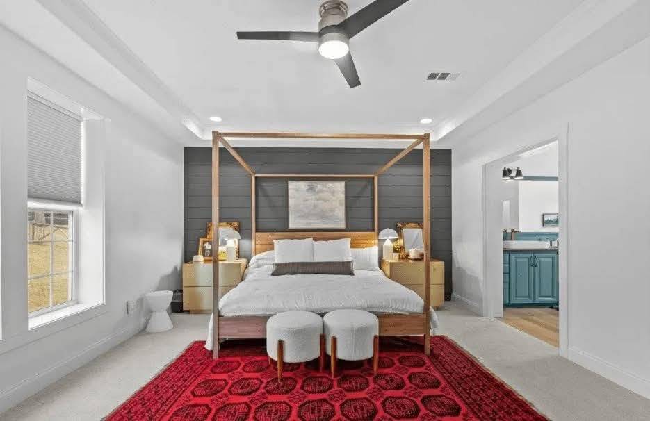 Modern bedroom with a canopy bed, white and gray bedding, flanked by wooden nightstands. A red rug adds warmth, and there's a visible bathroom.