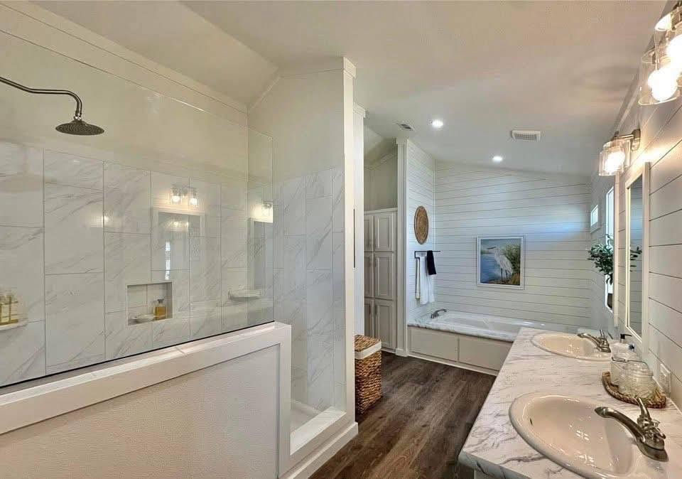 Elegant bathroom with a glass-enclosed shower, dual sinks on a marble counter, and a soaking tub. Wood flooring and soft lighting create a serene ambiance.