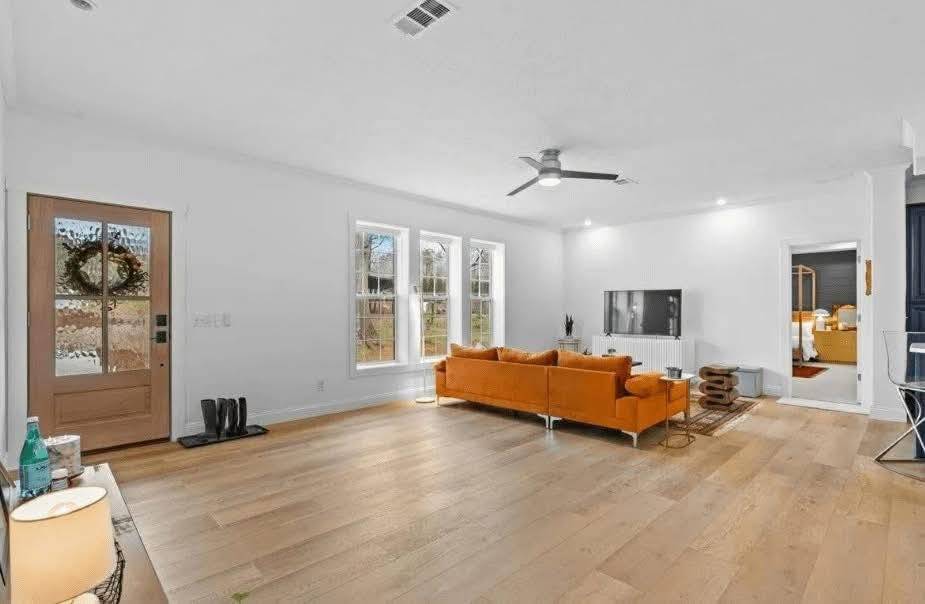 Spacious living room with a large orange sofa, wooden floor, and white walls. A ceiling fan hangs above, and natural light filters through three big windows.