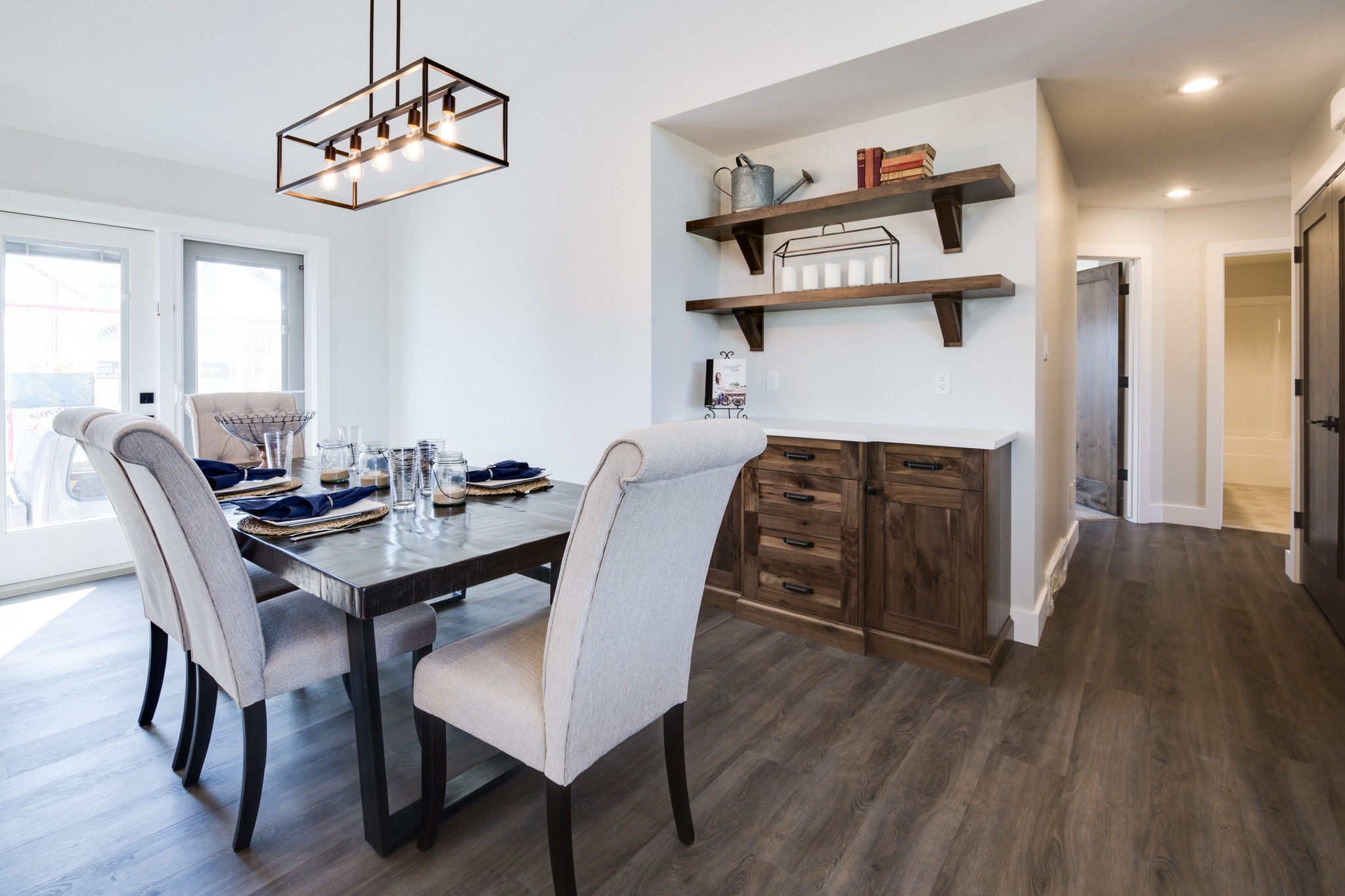 A modern dining room with a rectangular table set for four, featuring gray cushioned chairs. Wooden shelves and cabinets accent a cozy ambiance.