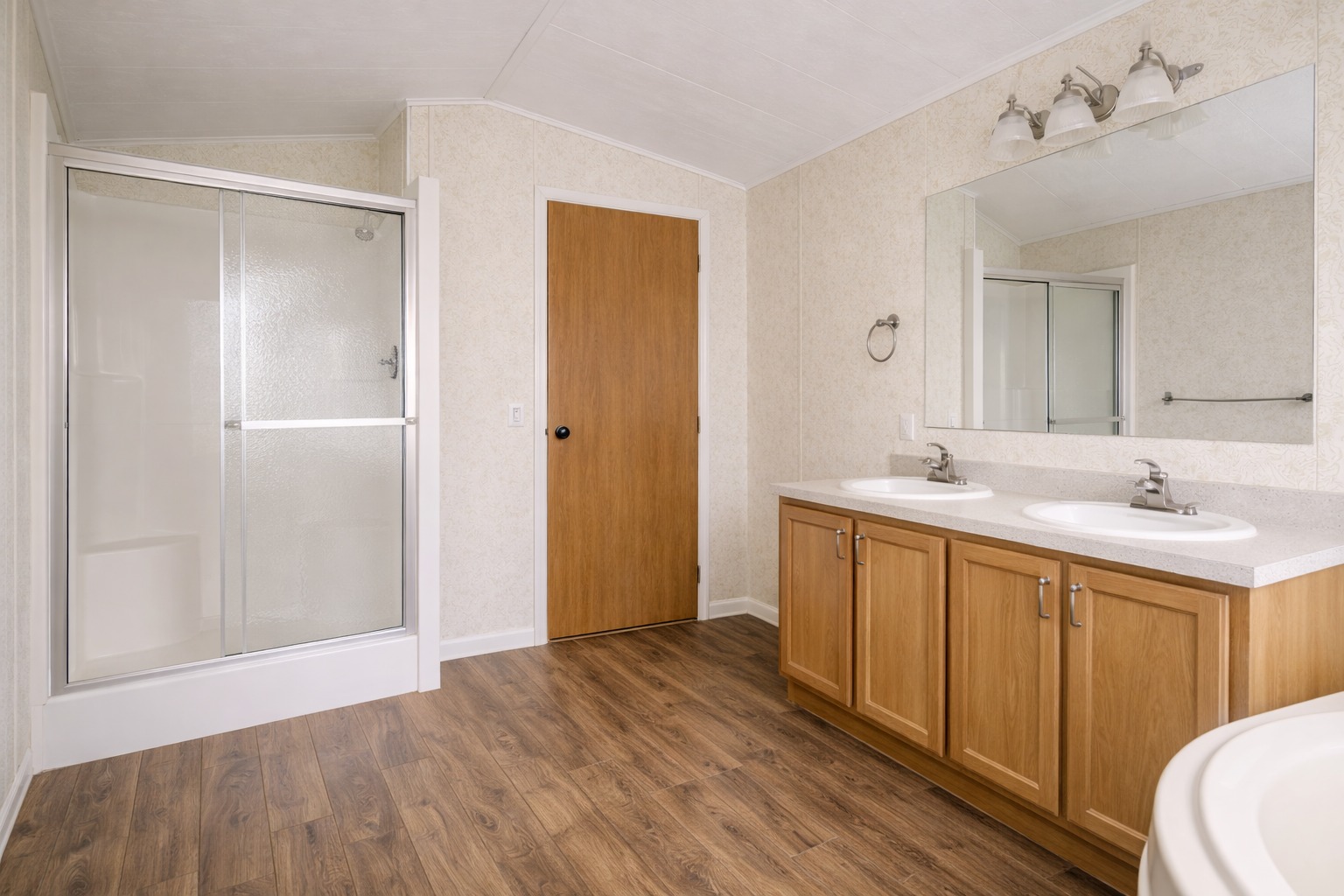 Spacious modern bathroom with wood floors, double sinks in wooden cabinets, a large mirror, glass shower, and wooden door. Clean and minimalist.