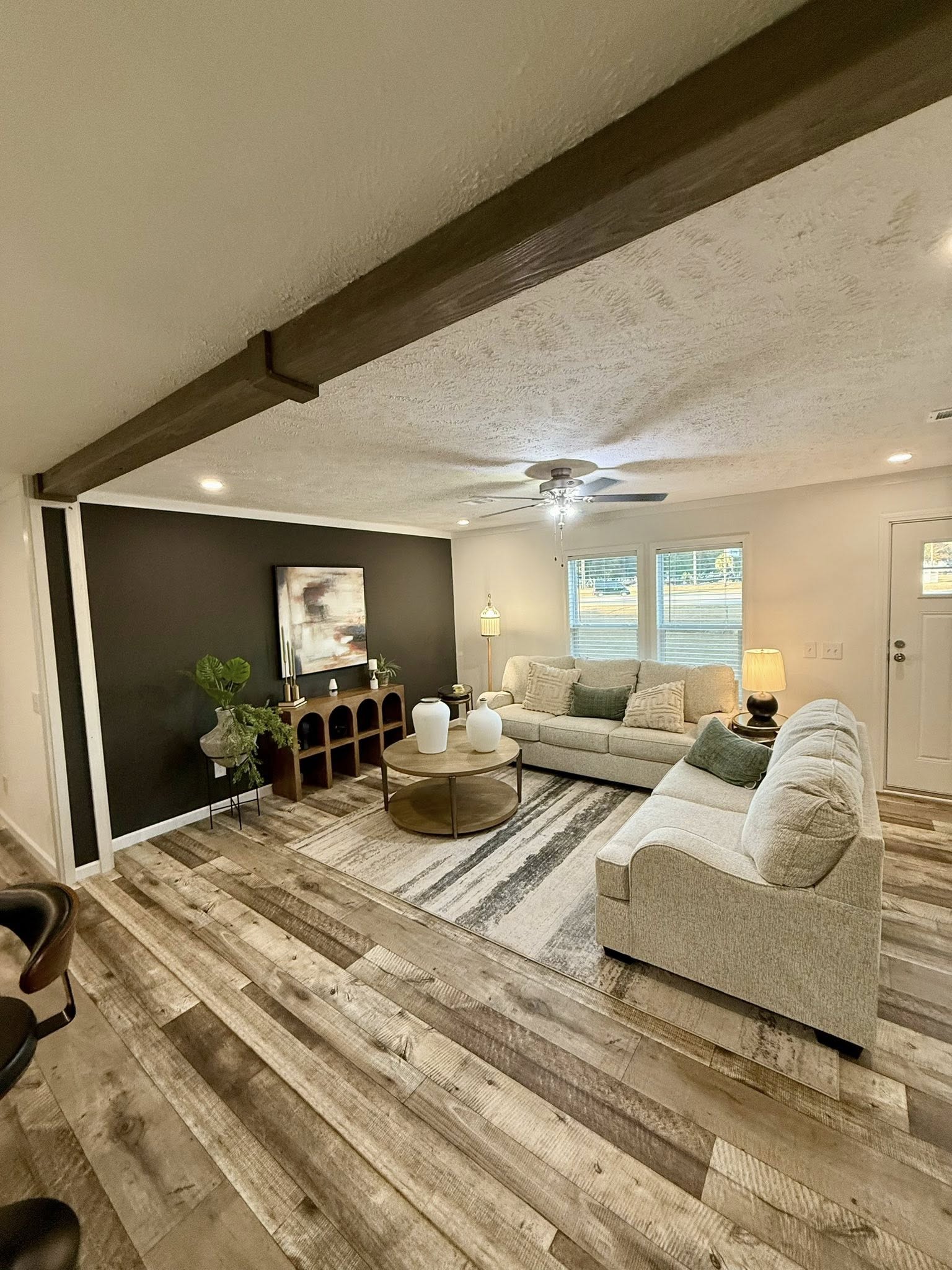 A cozy living room with light wood floors, two beige sofas, and a round coffee table. A dark accent wall features art and plants. Soft lighting and calm tones.