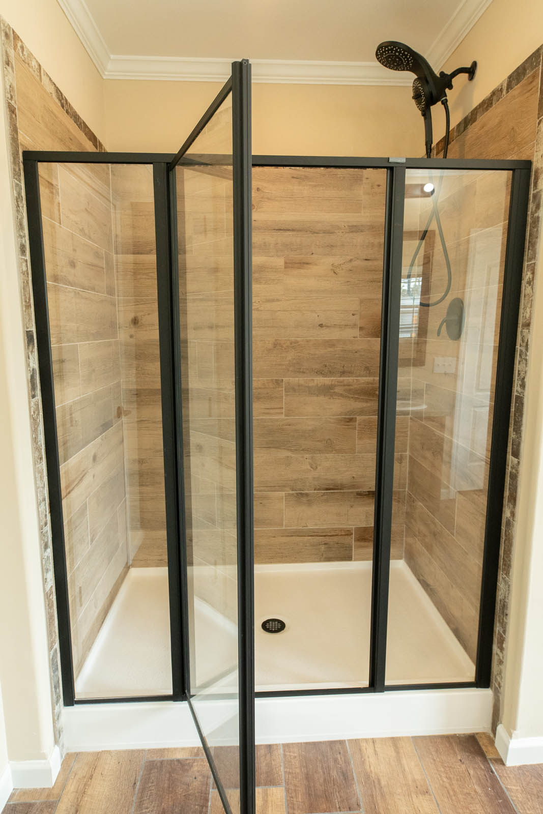A modern shower with black metal frames and glass doors. It features wood-textured tiles, a black showerhead, and a bright, clean atmosphere.