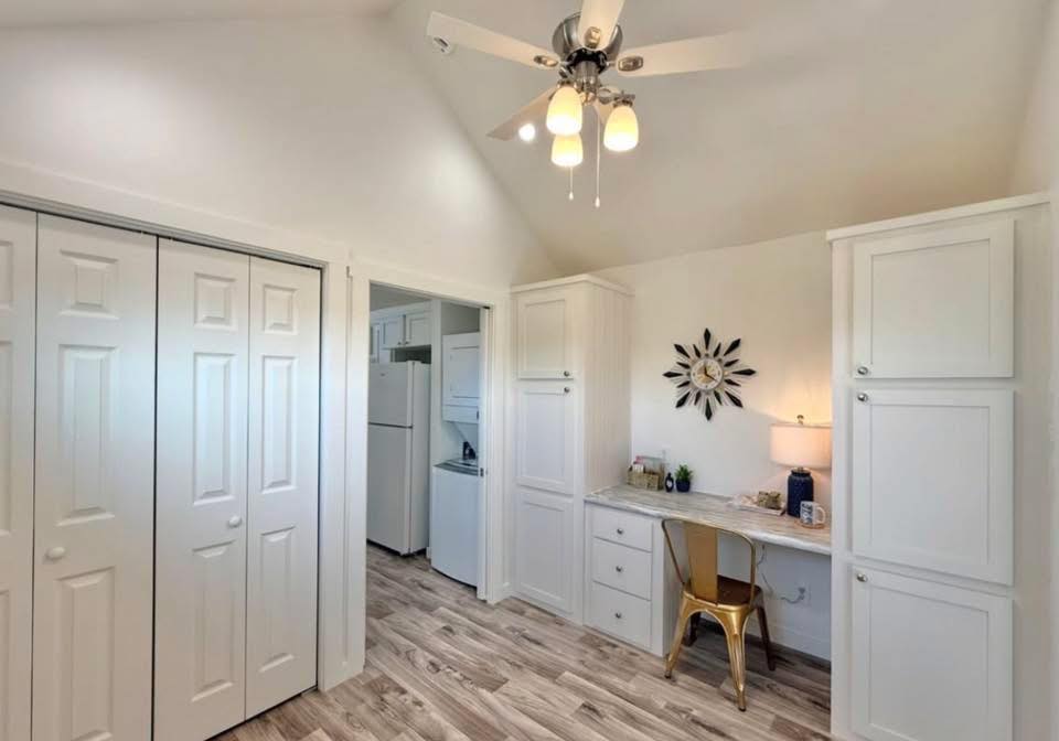 Bright room with white cabinets, a desk, and a chair. A ceiling fan with lights hangs above, and a sunburst wall decoration adds a modern touch.