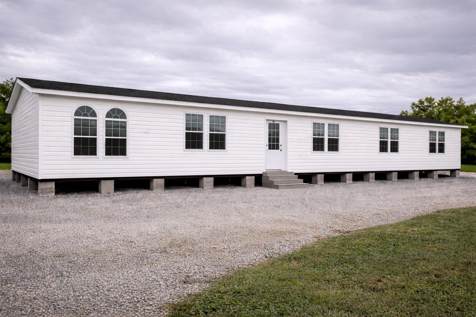 A white mobile home sits on a gravel lot under an overcast sky. It features multiple windows and a small central staircase. Green grass surrounds the area.