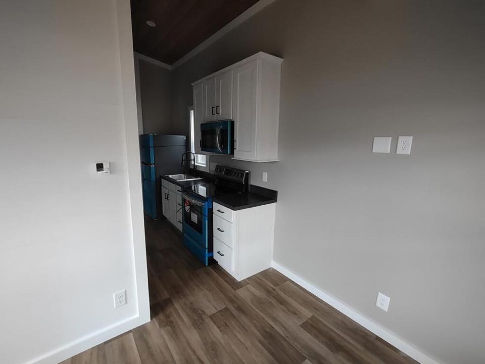 Compact kitchen with sleek white cabinets, black countertops, and stainless steel appliances, including an oven with blue accents. Wood flooring.
