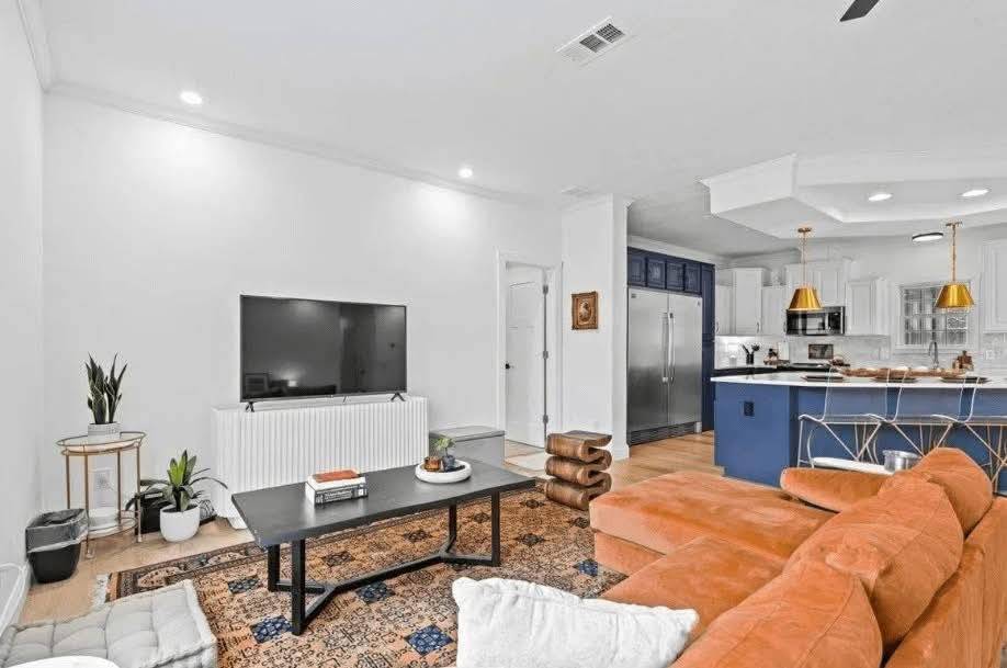 Modern living room with a sleek design. Rust-colored couch faces a TV on a white wall. A dark coffee table sits on a patterned rug. Blue and white kitchen with gold pendant lights in the background.