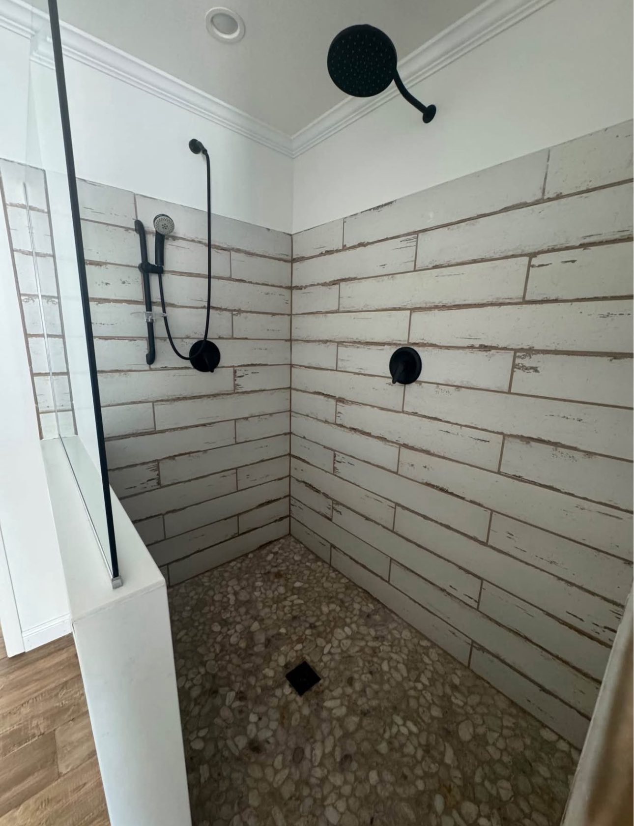 A modern shower with textured white tiles and pebble flooring. Black rain showerhead and hand shower are mounted on the walls, creating a sleek, minimalist look.