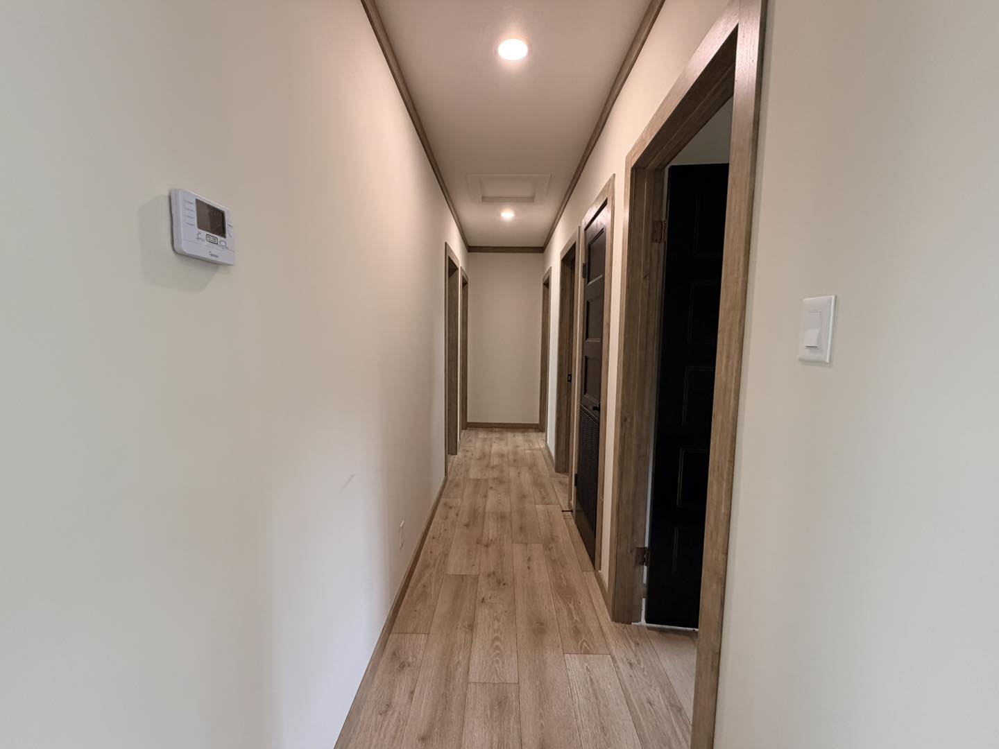 A narrow hallway with light wood flooring and beige walls leads to multiple wooden doorways. A thermostat is mounted on the left wall, and the space is brightly lit.