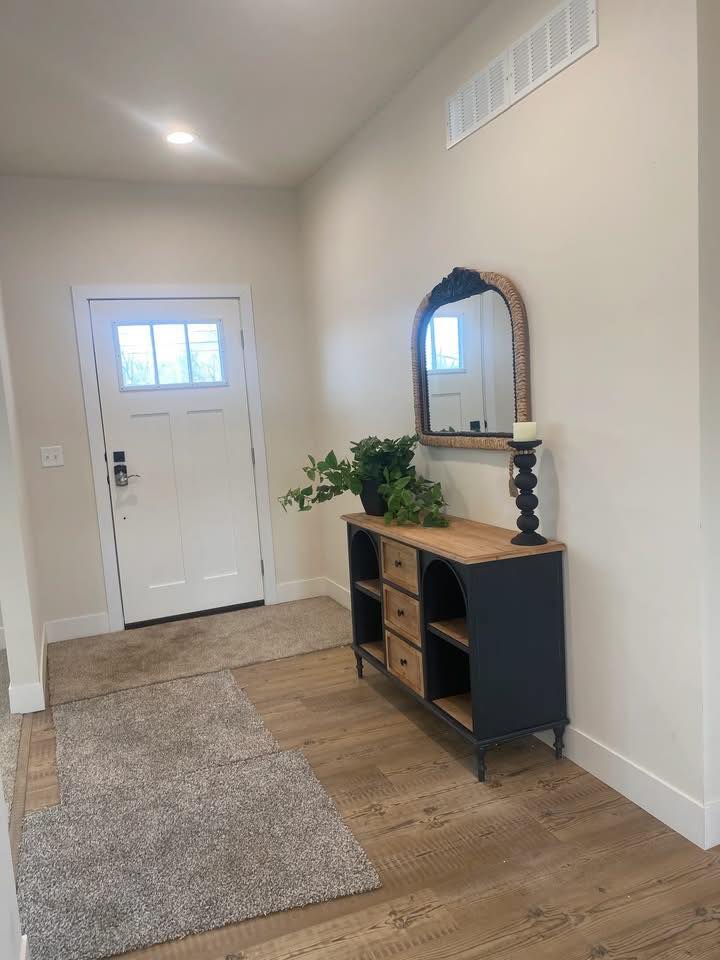 Warm-toned entryway with wood flooring, white door, and a black side table. A mirror with a wicker frame hangs above, with greenery and a candle adding cozy touches.