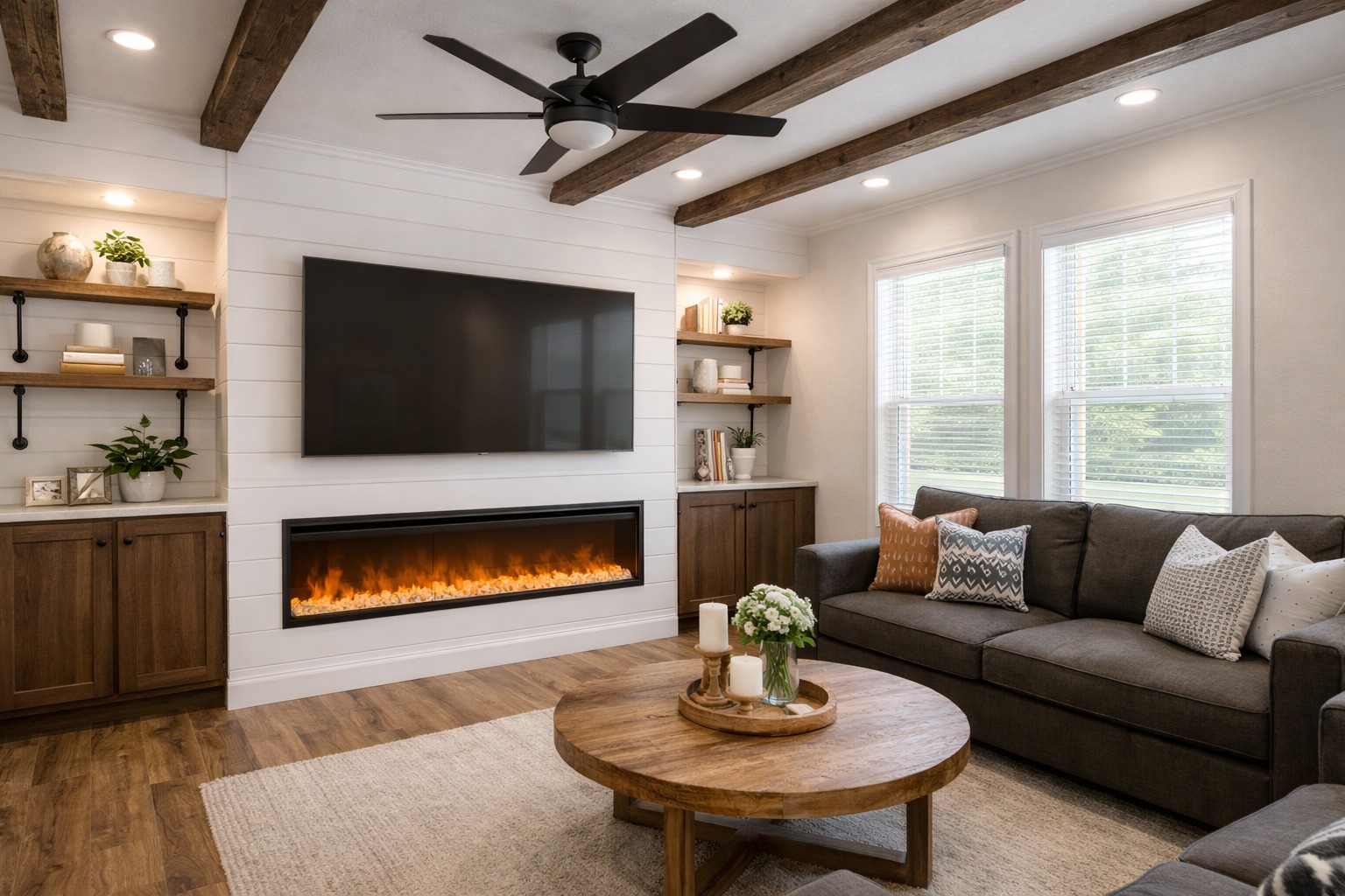 A cozy living room with a modern design features a mounted TV above a sleek electric fireplace. A dark sofa, wooden shelves, and beams add warmth.