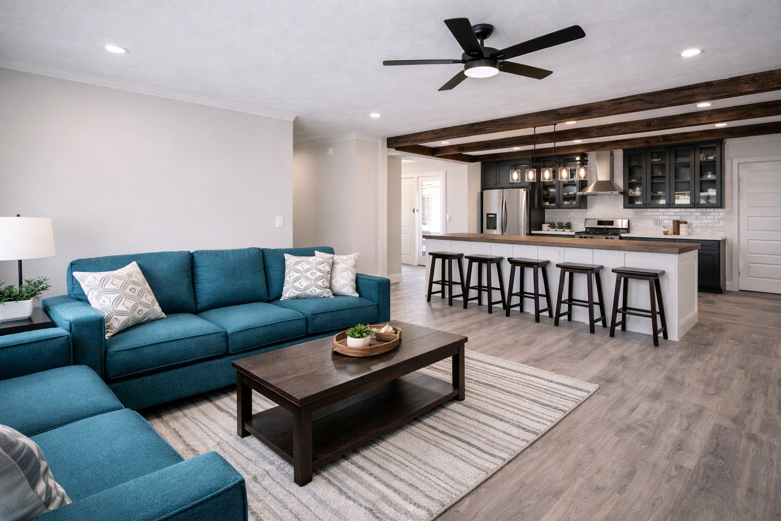 Modern living room with a teal sofa, patterned pillows, and a dark wood coffee table on a striped rug. Open kitchen with bar stools and exposed beams. Cozy and inviting.