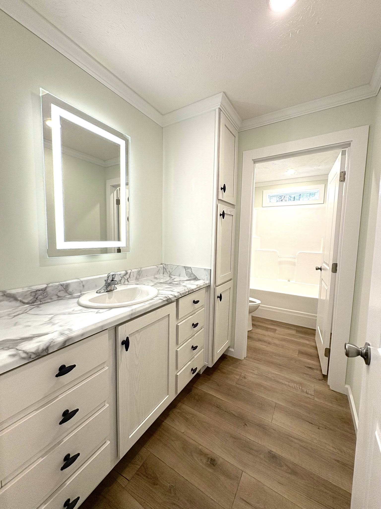 Bright bathroom with wood floor, marbled countertop, white cabinets, and lit rectangular mirror. Doorway leads to a shower-tub combo. Calm and modern feel.