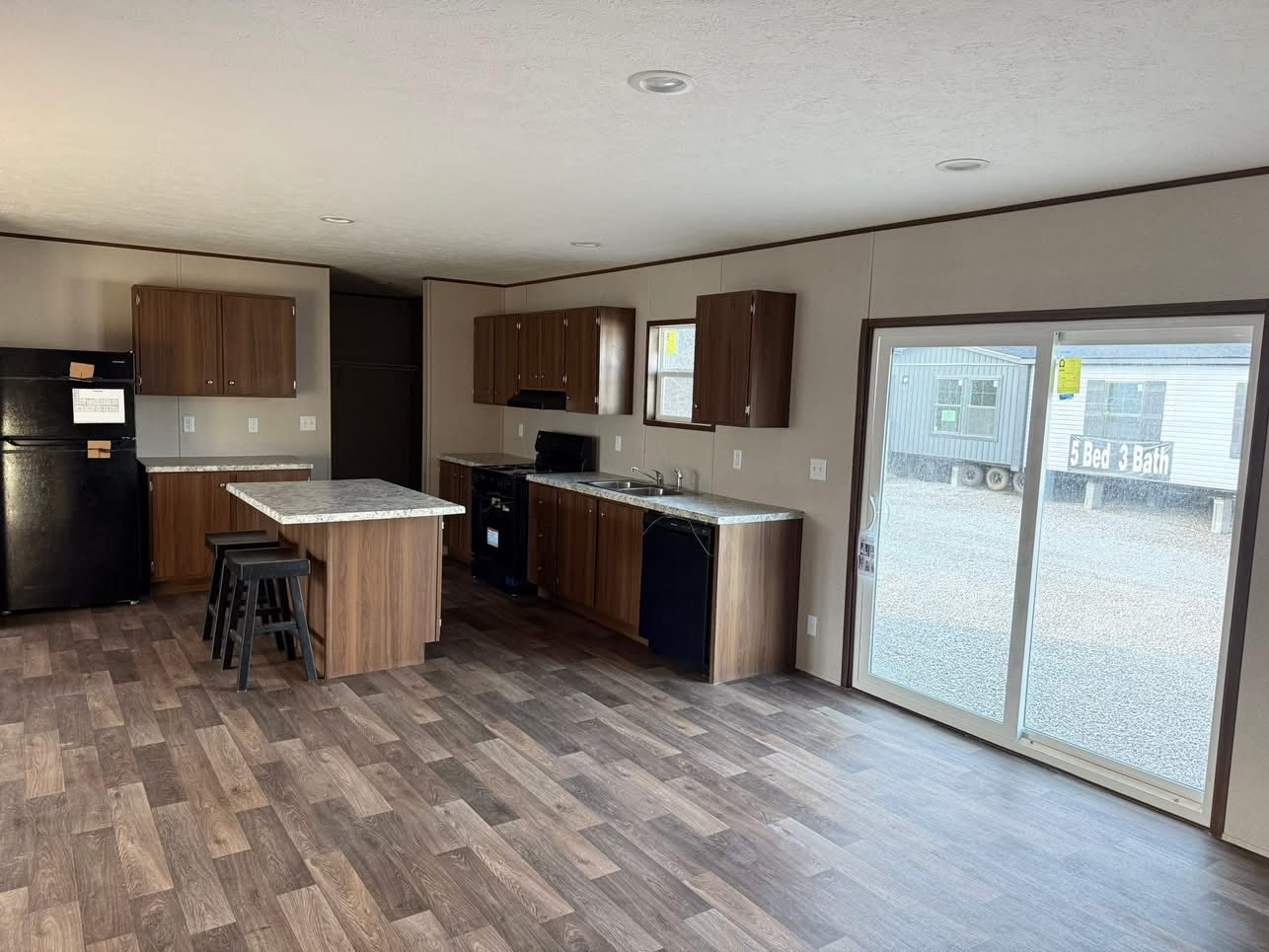 Spacious kitchen with dark wood cabinets, island with stools, black appliances, and sliding glass door. Laminate wood floor adds warmth.