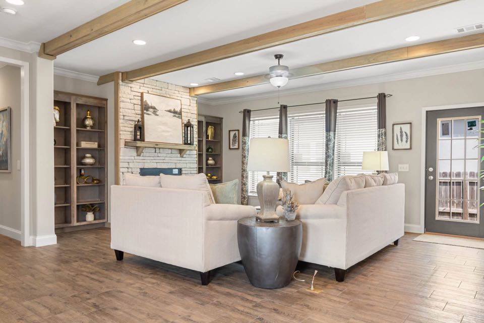Cozy living room with beige sectional sofa, stone fireplace, wooden shelves, and large windows. Warm lighting creates a welcoming atmosphere.