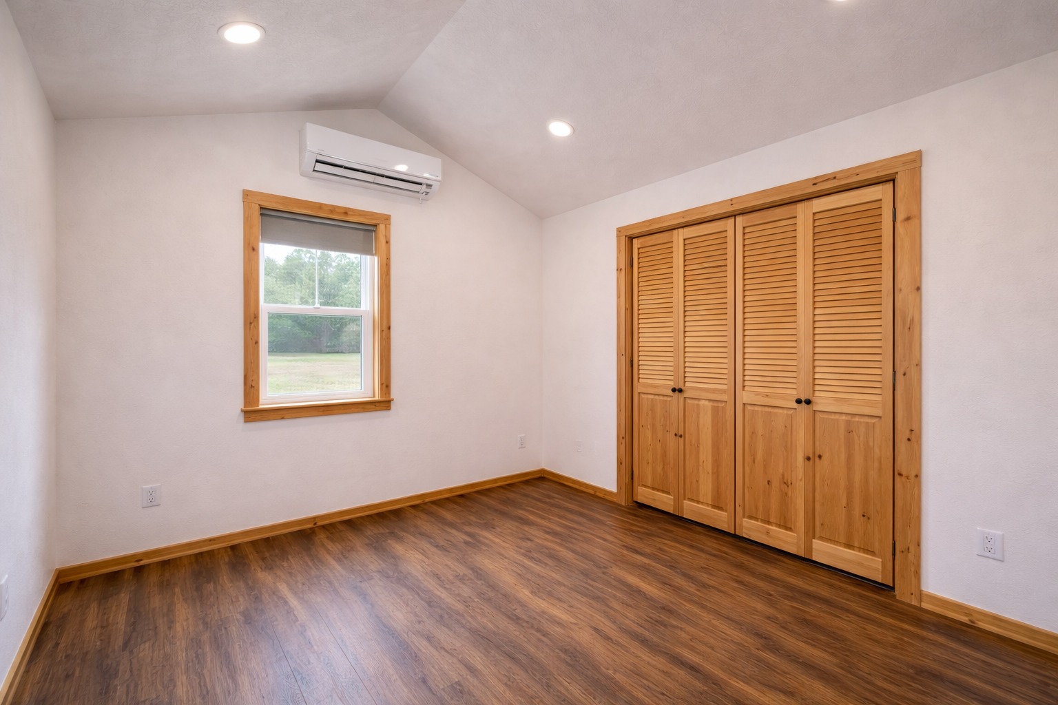 Empty room with wooden floor, beige walls, and recessed lighting. It features a window with wooden trim, an AC unit above, and a double louvered closet.
