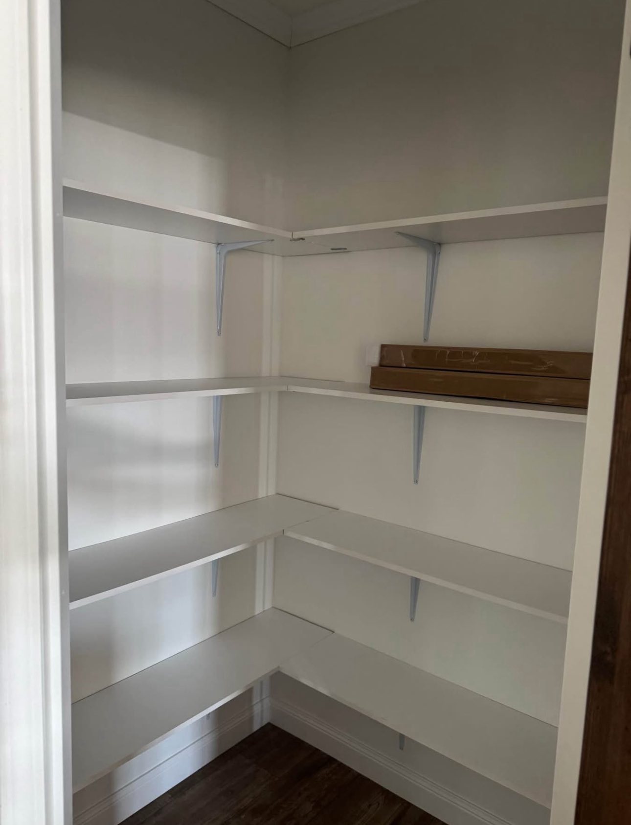 Empty pantry with white shelves and a wooden floor. Cardboard box on the top shelf. The space appears neat and ready for organizing.