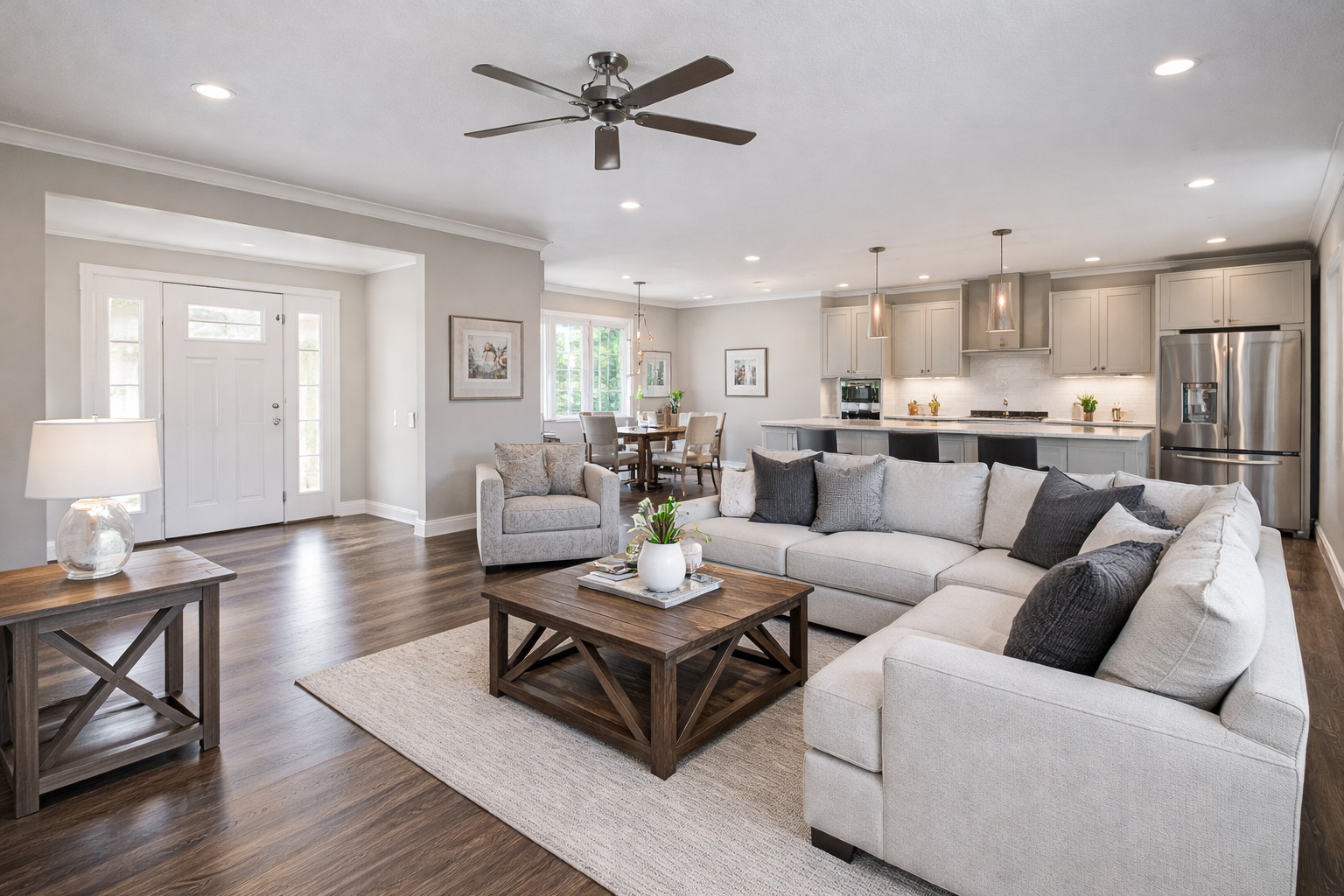 Modern open-concept living area with light gray sofas, wooden coffee table, and kitchen in the background. Neutral tones, natural light, cozy atmosphere.