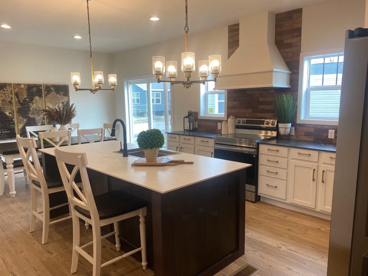A cozy kitchen with a central island, white cabinets, wood flooring, and brick accents. A chandelier hangs above the island, creating a warm ambiance.