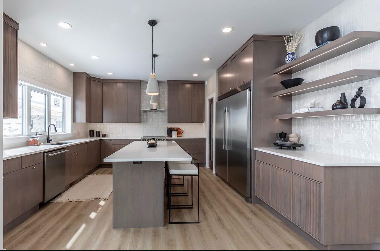 Modern kitchen with sleek wooden cabinets and white countertops. An island with stools is centered, flanked by stainless steel appliances and open shelves. The room is bright with natural and recessed lighting, evoking a clean, minimalist vibe.