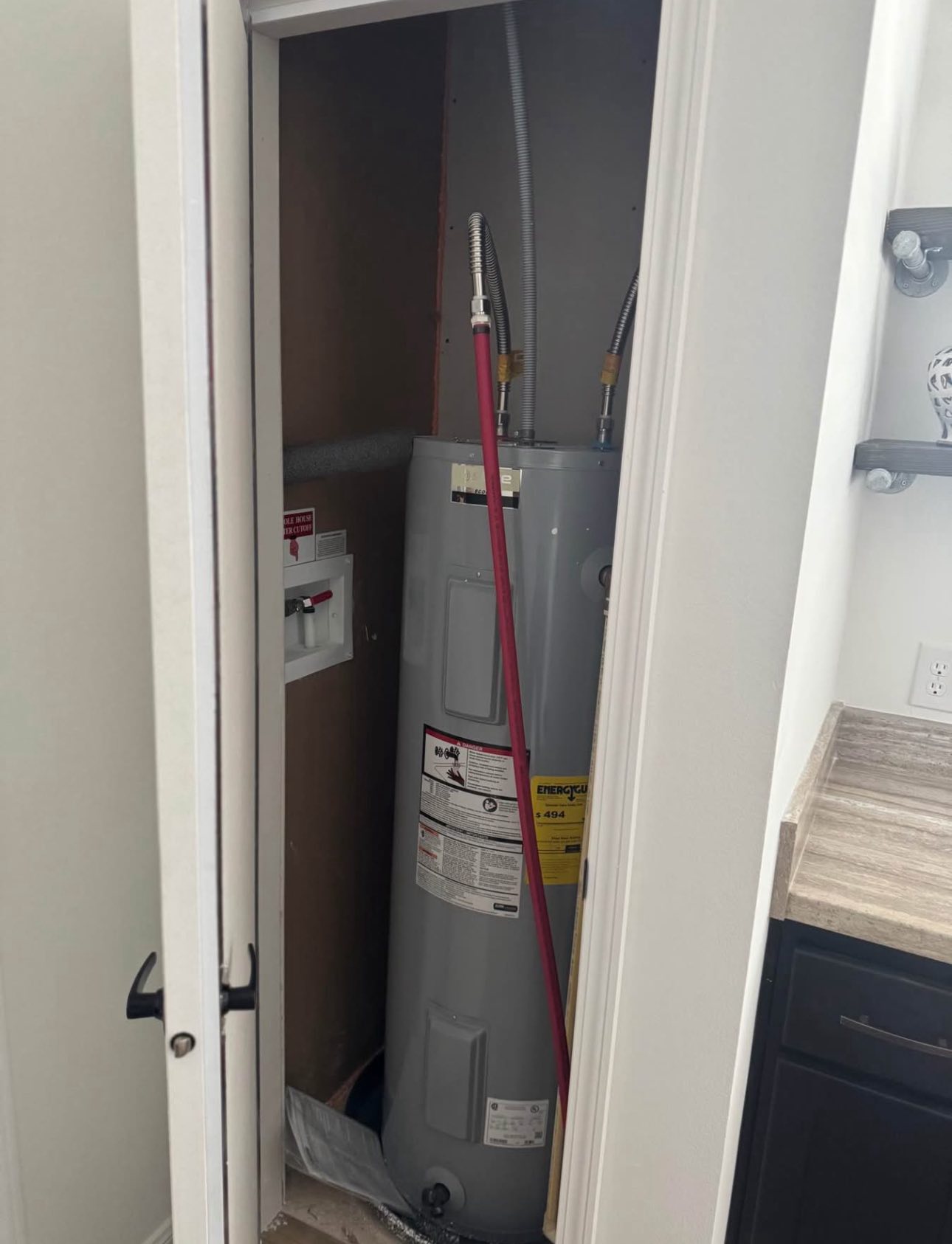 A water heater in a small closet with pipes connected, a red handle nearby, next to a countertop. The scene is practical and functional.