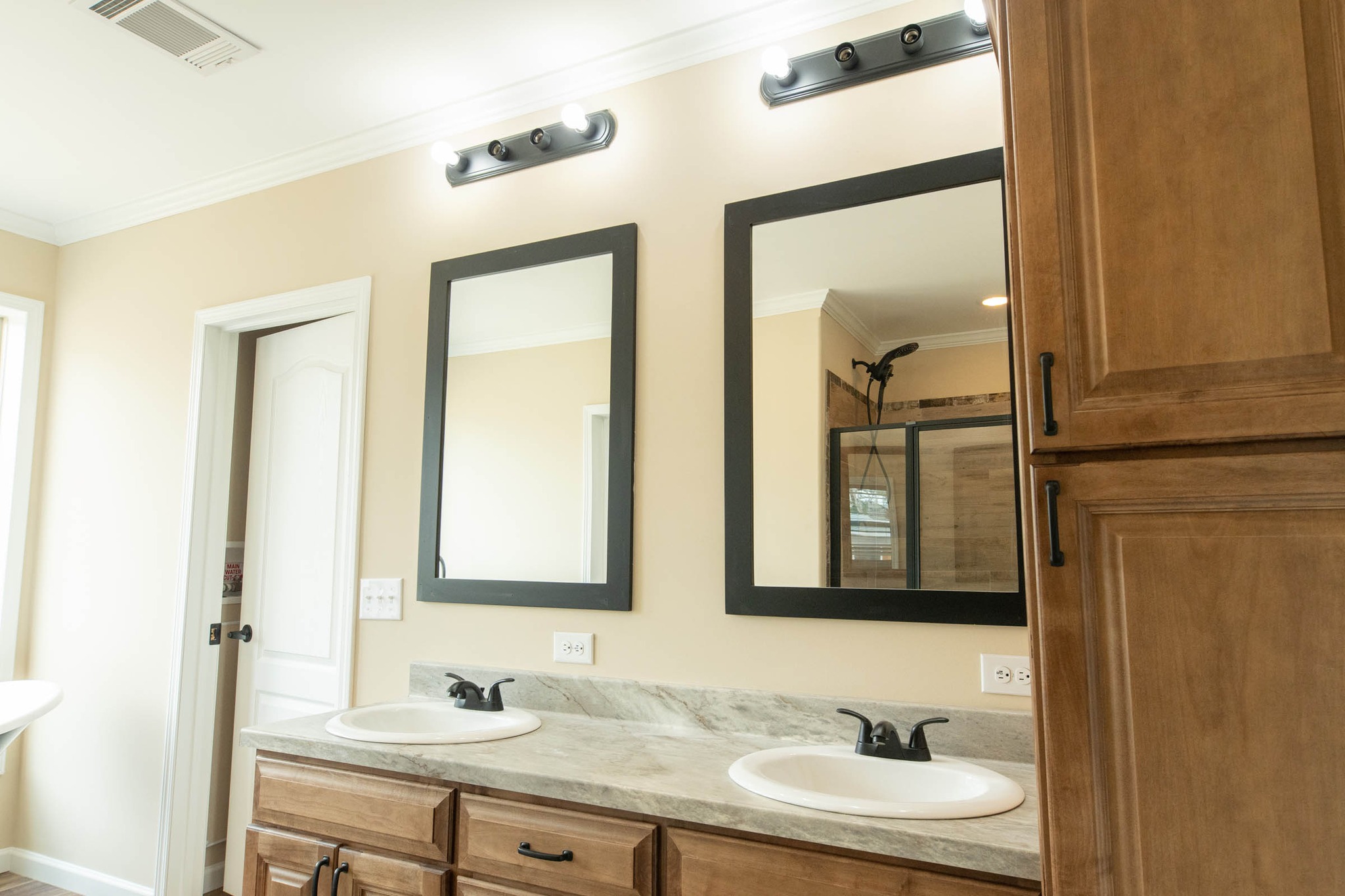 A modern bathroom features dual sinks with black faucets, each under rectangular mirrors. Light fixtures above are on, highlighting beige walls and wood cabinetry.