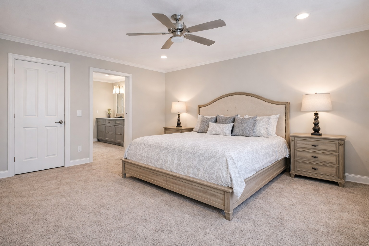 Spacious bedroom with a large bed, beige bedding, and two wooden nightstands with lamps. There's a ceiling fan and a doorway leading to a bathroom.