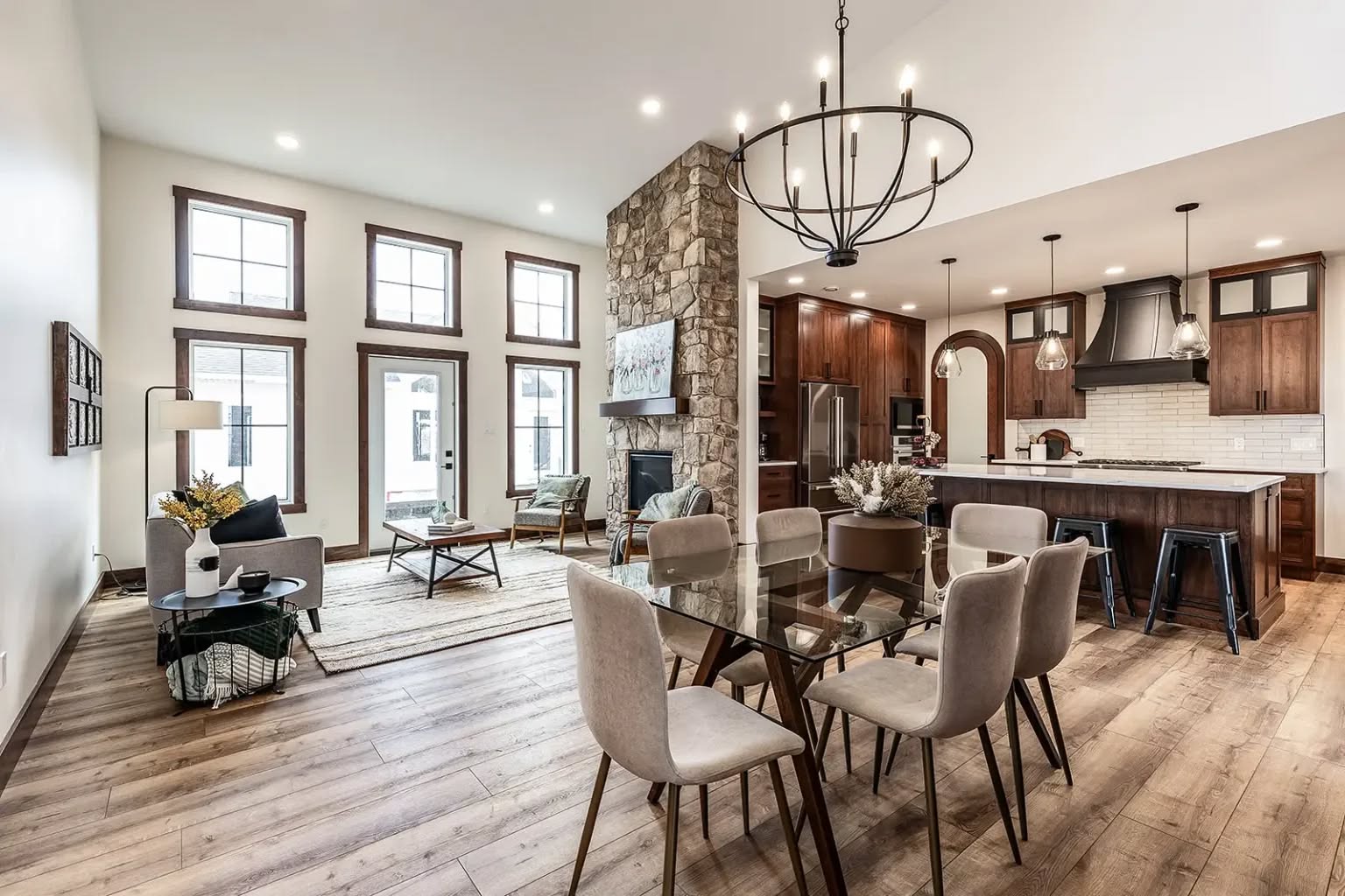 Spacious open-plan living area with stone fireplace, modern chandelier, and glass dining table. Warm tones, wooden floors, and large windows create an inviting atmosphere.