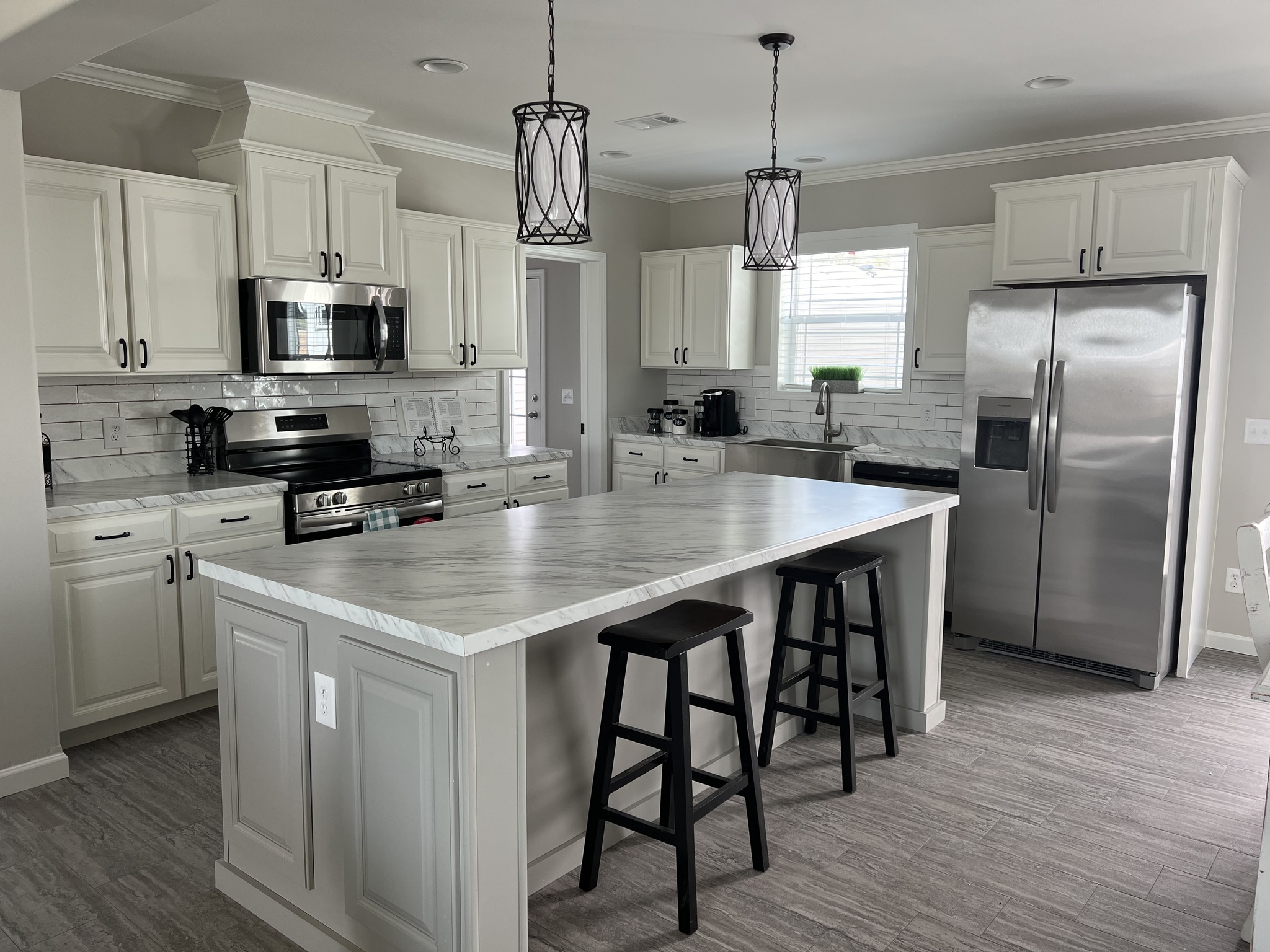 Spacious kitchen with white cabinets, stainless steel appliances, large marble island with two black stools, pendant lights, and gray tile flooring.