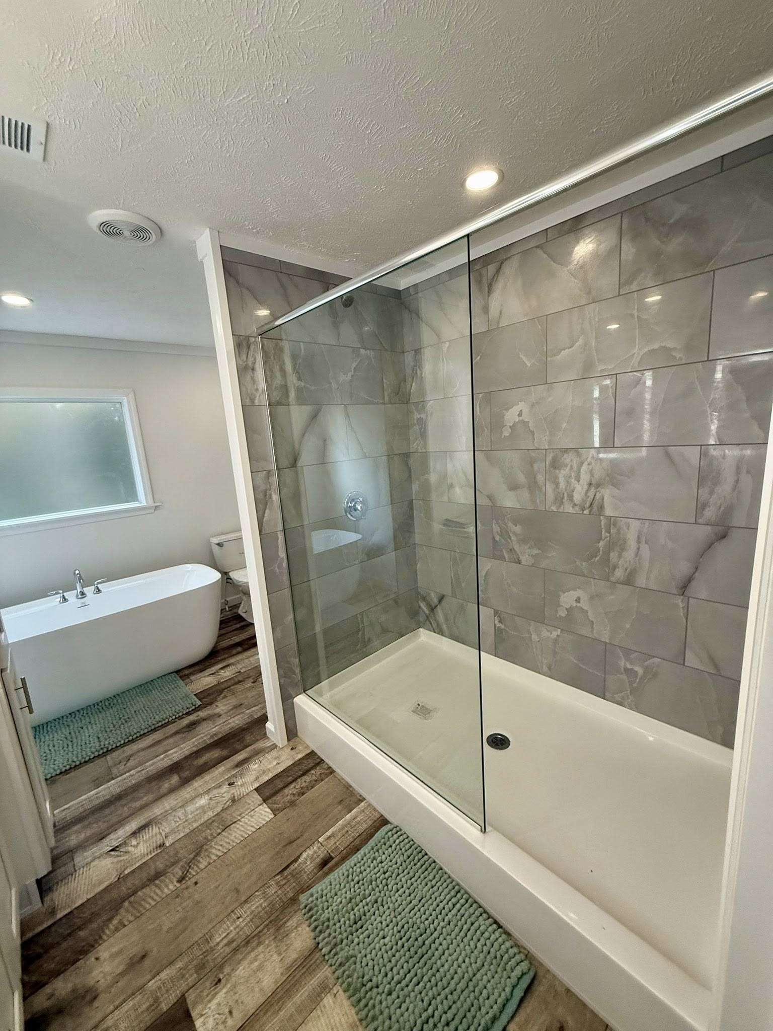 Modern bathroom with double sinks, lit mirrors, and wood flooring. A freestanding bathtub with a green mat is visible. Calm and elegant ambiance.
