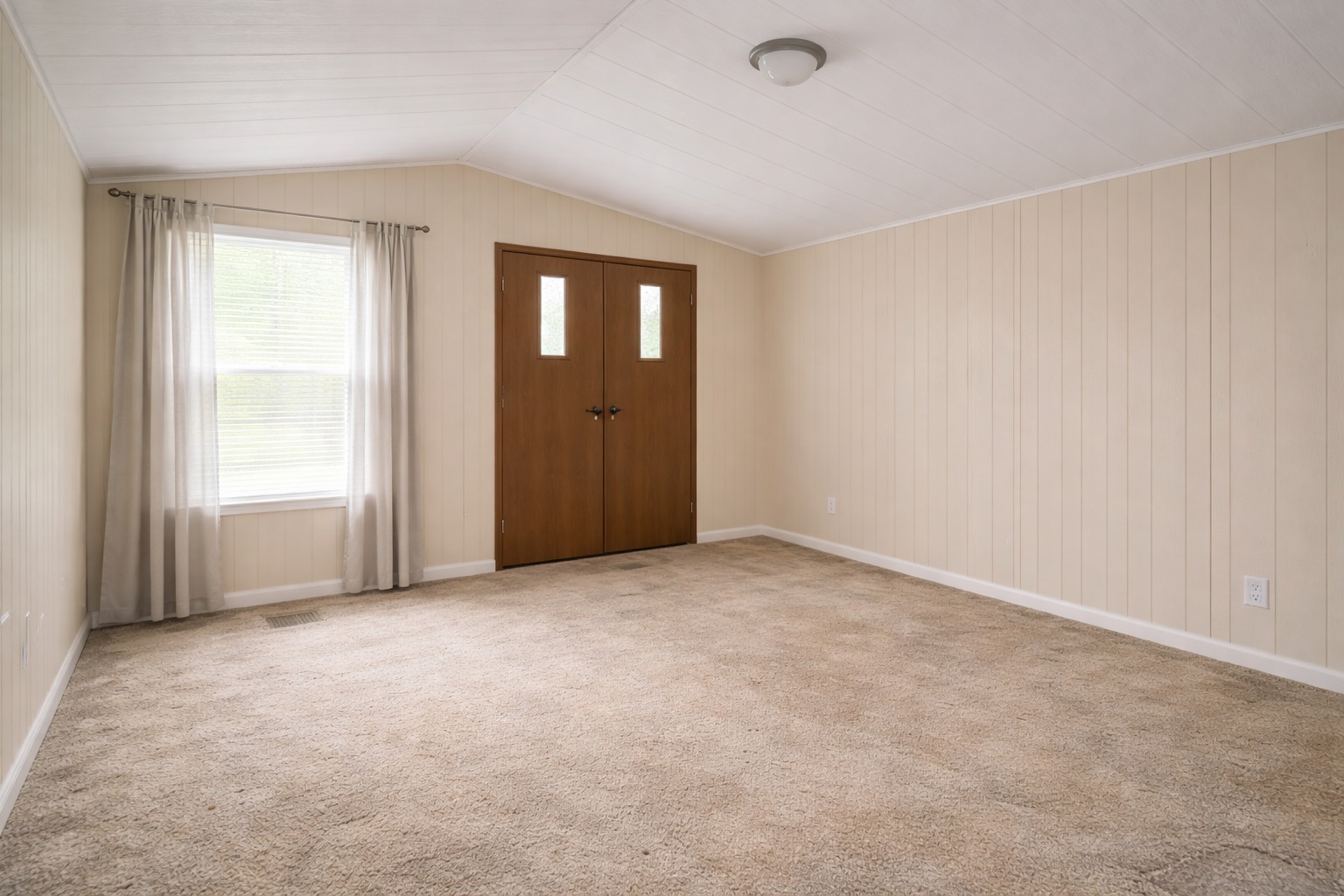 Empty room with beige carpet, light cream wood-paneled walls, a large window with sheer curtains, and wooden double doors. The space feels open and neutral.