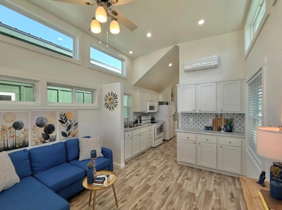 Bright modern living room with vaulted ceiling, featuring a blue sectional sofa, abstract art, small round table, and adjacent kitchen with white cabinets.