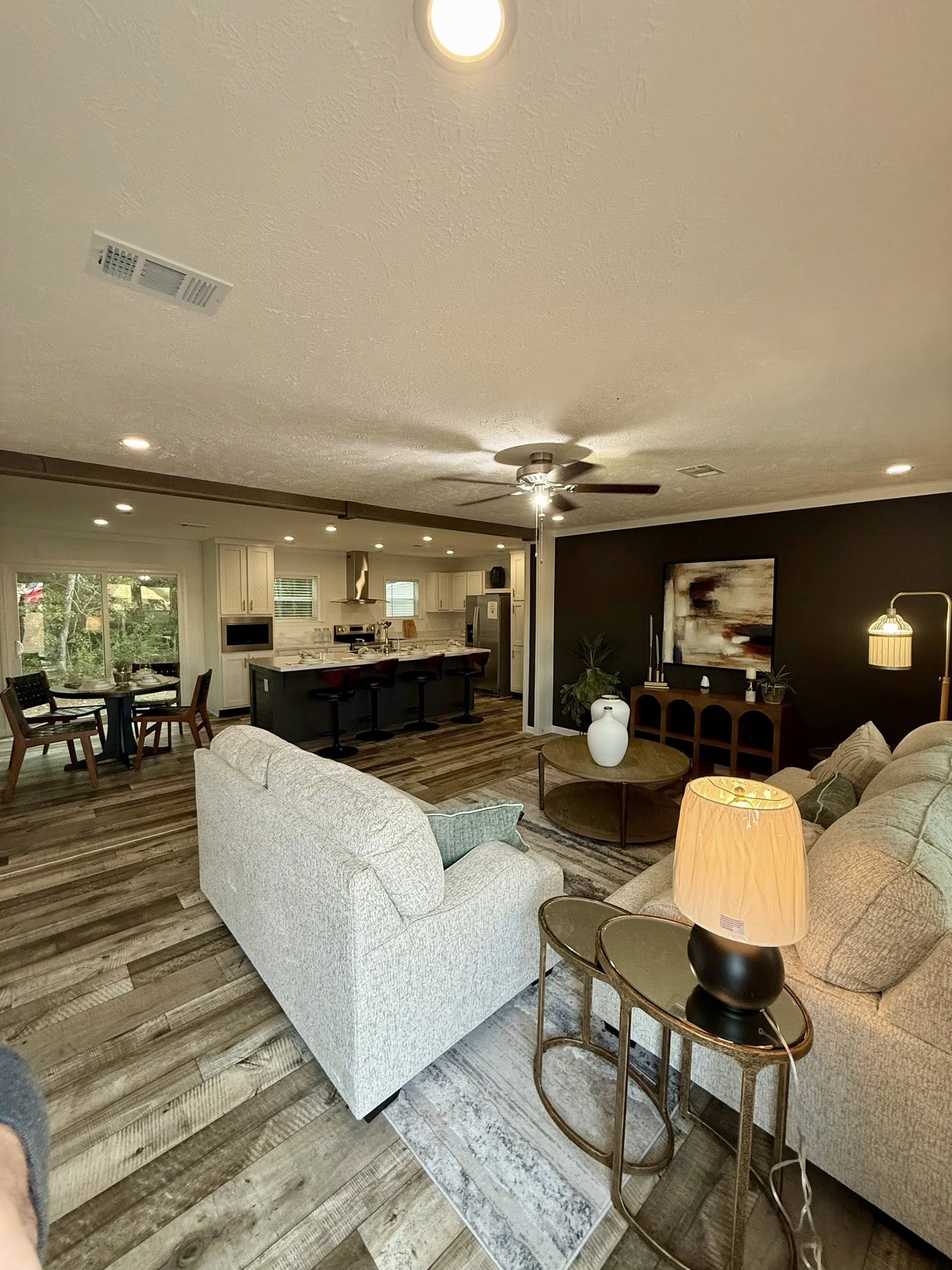 Open-concept living room with rustic wood flooring, cozy beige couches, and a black accent wall featuring abstract art. Nearby, a kitchen island has bar stools, and a dining area overlooks a garden. Warm lighting adds a welcoming ambiance.
