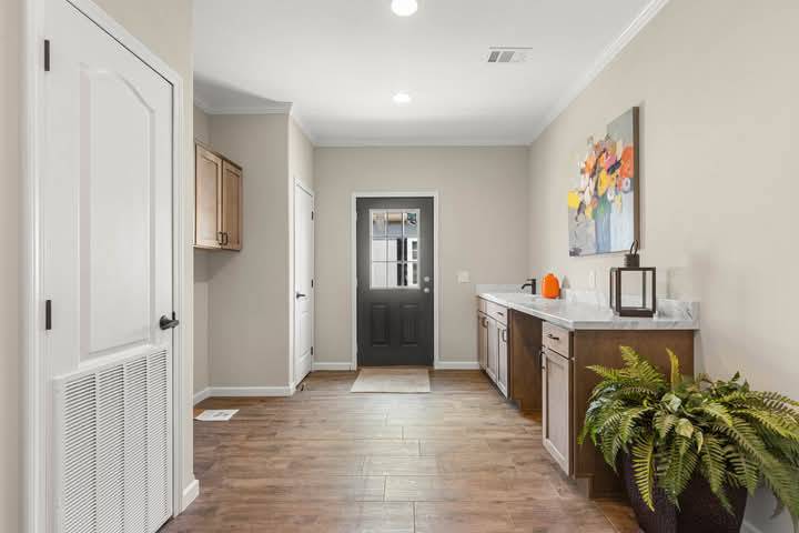 A hallway with wooden flooring and neutral walls features a dark door at the end. On the right, there's a countertop with a small pumpkin, lantern, and vibrant floral art. A potted plant sits below, adding a touch of greenery. A wood cabinet is on the left. The space feels inviting and orderly.