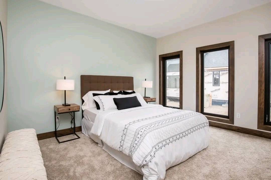 Modern bedroom with light green accent wall, king-size bed with white and patterned linens, two wooden nightstands, lamps, and three tall windows. Cozy ambiance.