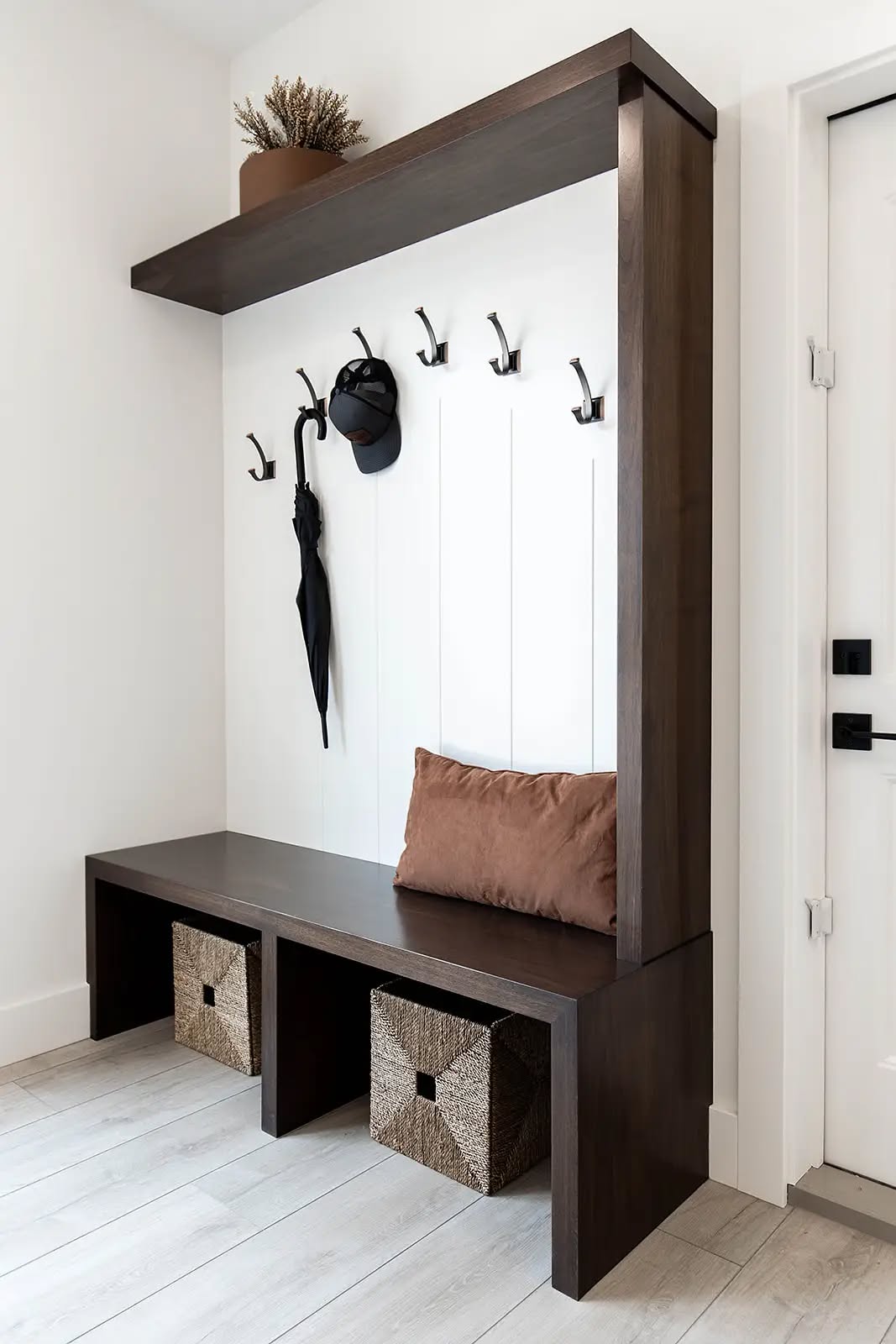 Modern entryway with a dark wood bench and coat rack. Hooks hold a black cap and umbrella. Brown cushion on bench; two woven baskets below. Minimalist style.