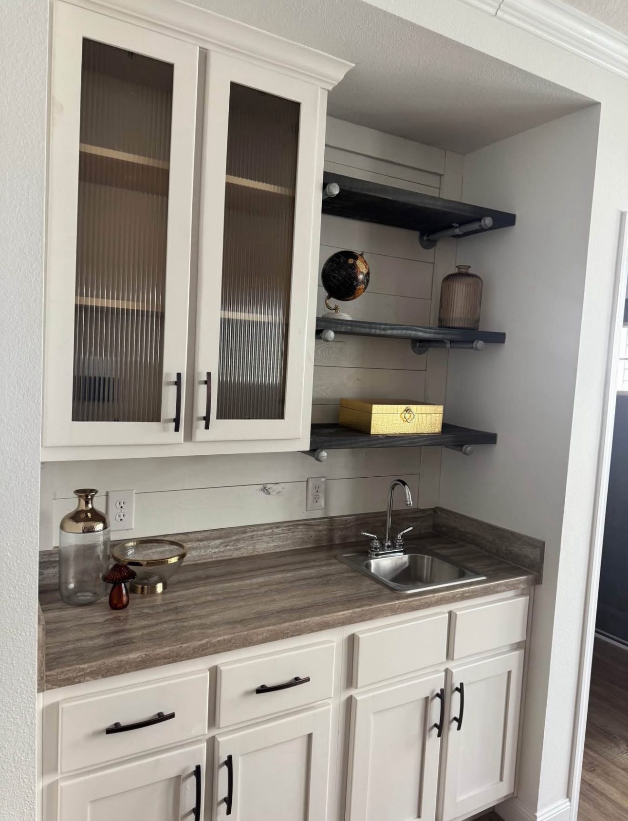 Small kitchen nook with white cabinets, glass-paneled doors, and a gray countertop. Open shelves hold a globe, basket, and jar. Sink on the right.
