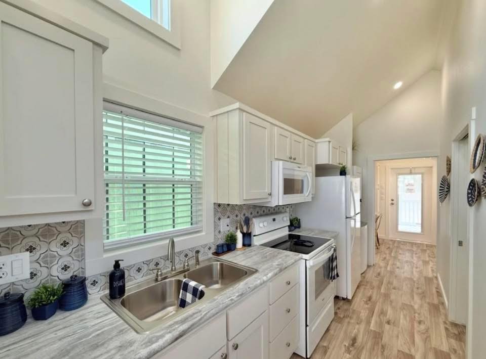 Bright, narrow kitchen with white cabinets and wood flooring. A large window above a double sink, modern appliances, and decorative tiles add charm.