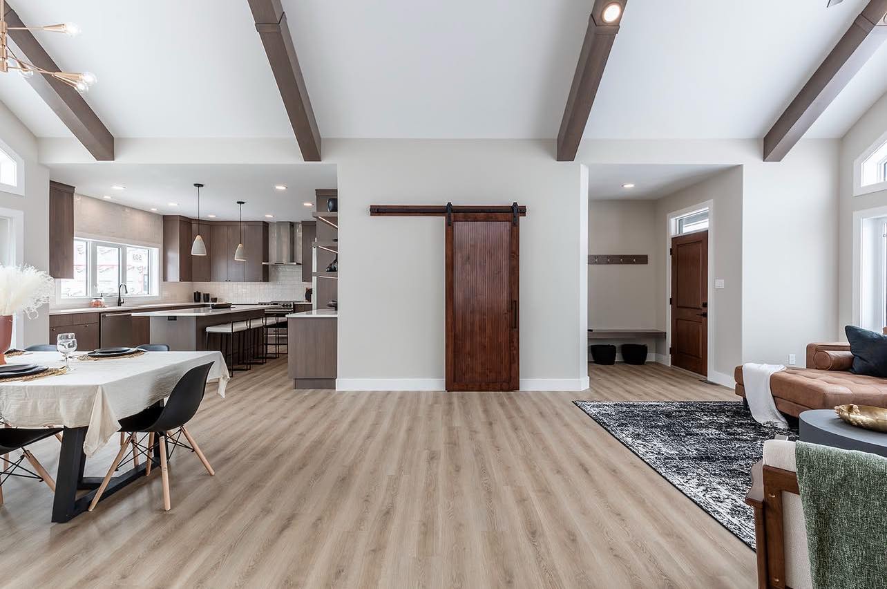 Spacious modern living area with light wood flooring, exposed beams, and a sliding barn door. Features a cozy kitchen, dining table, and stylish decor.